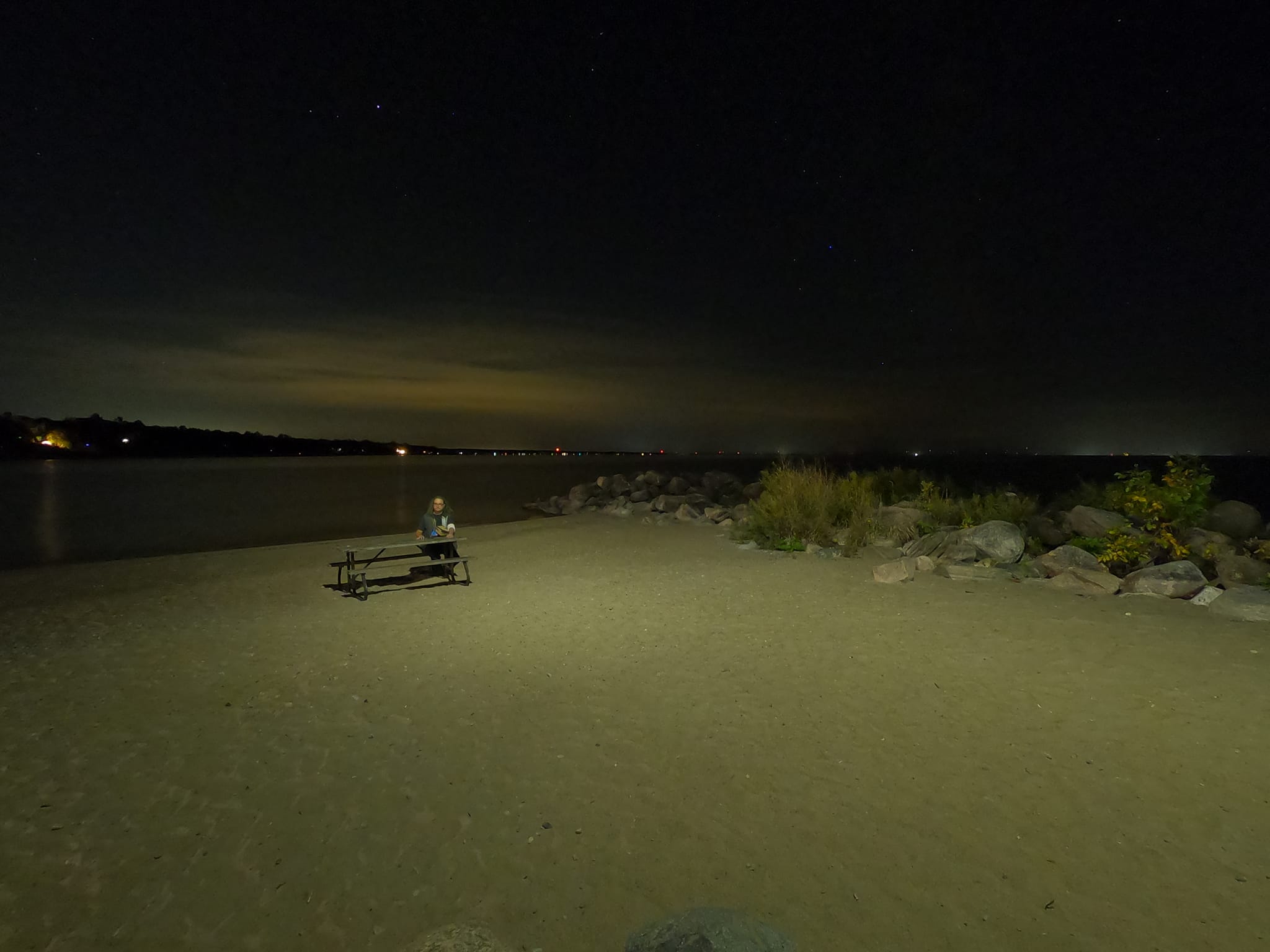 A man with long green hair and no beard sits at a picnic table on a beach with water in the background"